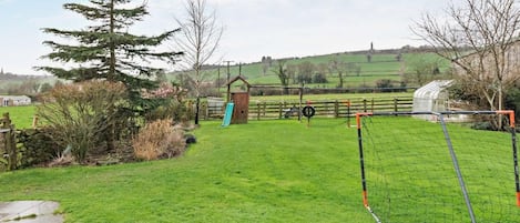 A grassy outdoor area with a small wooden structure, a fenced play area, and a stone wall.