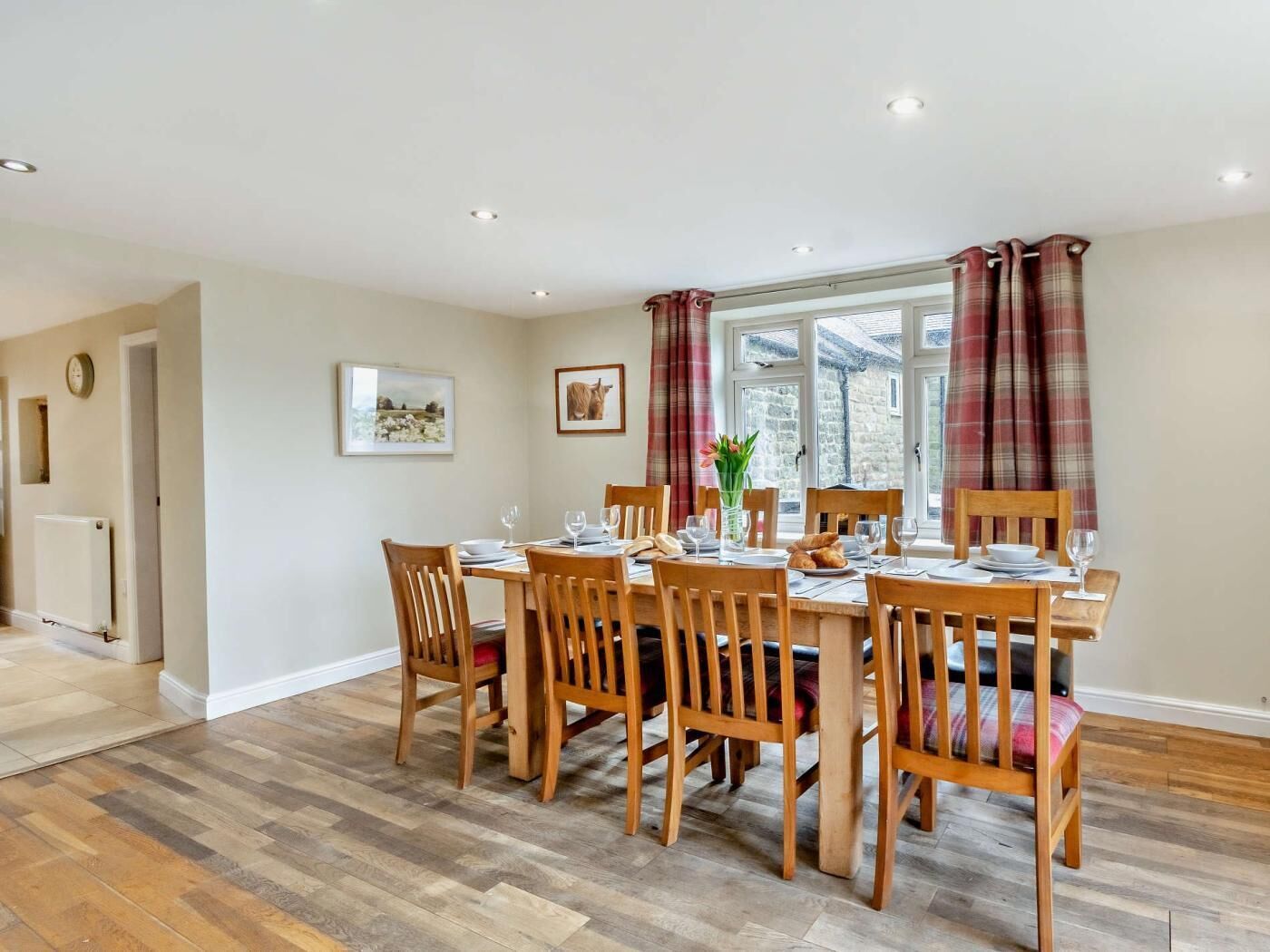 A dining room with a wooden table set for six, flanked by two windows with plaid curtains, a clock on the wall, and a framed picture above the table.