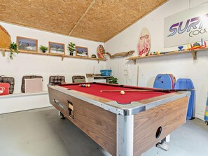 Ett biljardbord i ett rum med ett träpanelat tak, en skylt med en surfbräda och olika inramade tavlor på väggen.