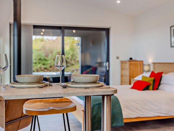 A modern room with a dining table set for two, a bed with colorful pillows, and a view through a glass door.