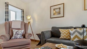 Ein Wohnzimmer mit einem gemusterten Sessel, einem grauen Sofa, einer Stehlampe, einem gerahmten Bild, einer Zeitschrift, einer Kaffeetasse und einem Teekessel auf einem Holztisch.