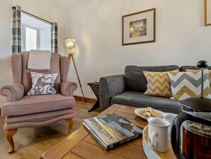 Uma sala de estar com uma poltrona estampada, um sofá cinza, um abajur, um quadro emoldurado, uma revista, uma xícara de café e uma chaleira sobre uma mesa de madeira.