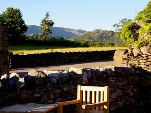 A stone wall with a wooden bench, a paved area, and a road leading to a mountainous landscape. - 1 Bed in Penrith (oc-94068) (Ullswater)