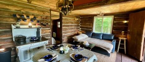 L’intérieur chaleureux d’un chalet comprend une table à manger pour deux personnes, un canapé et un coin cuisine avec un poêle.