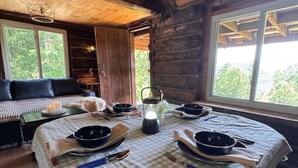 Interior acollidor d'una cabana amb taula de dinar preparada per a quatre, una làmpada de cristall i un gran finestral que ofereix vistes a l'exterior.
