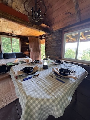 A cozy cabin interior with a dining table set for four, a chandelier, and a large window offering a view of the outdoors. - Cozy Rustic Cabin with Scenic Views (Johnsonville)