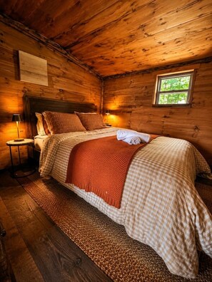 A wooden cabin bedroom with a bed, a nightstand, a lamp, and a window. - Cozy Rustic Cabin with Scenic Views (Johnsonville)