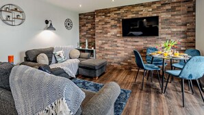 A modern living room with a brick wall, a flat-screen TV, a sofa, a coffee table, and a dining area with chairs. - 2 Bed in Cromer (oc-91297) (Cromer)