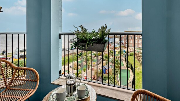 A balcony with wicker chairs, a table with a coffee pot and cups, and a hanging planter with green plants. - 2 Bed in Cromer (oc-91297) (Cromer)