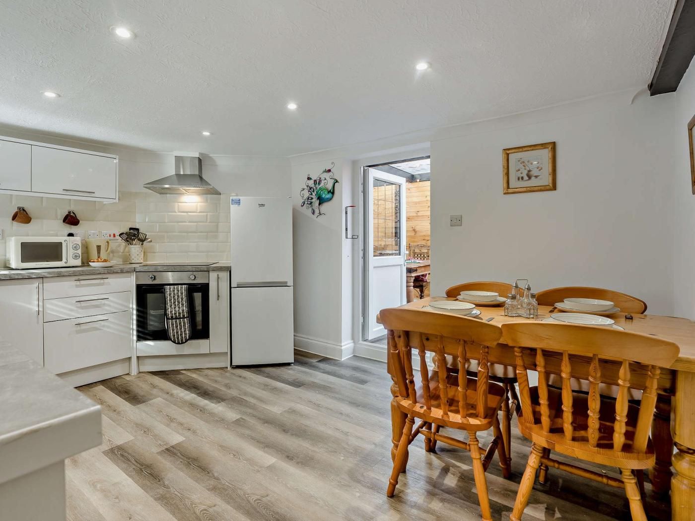 A modern kitchen with white cabinets, a built-in oven, and a refrigerator. There is a dining area with a wooden table and chairs.