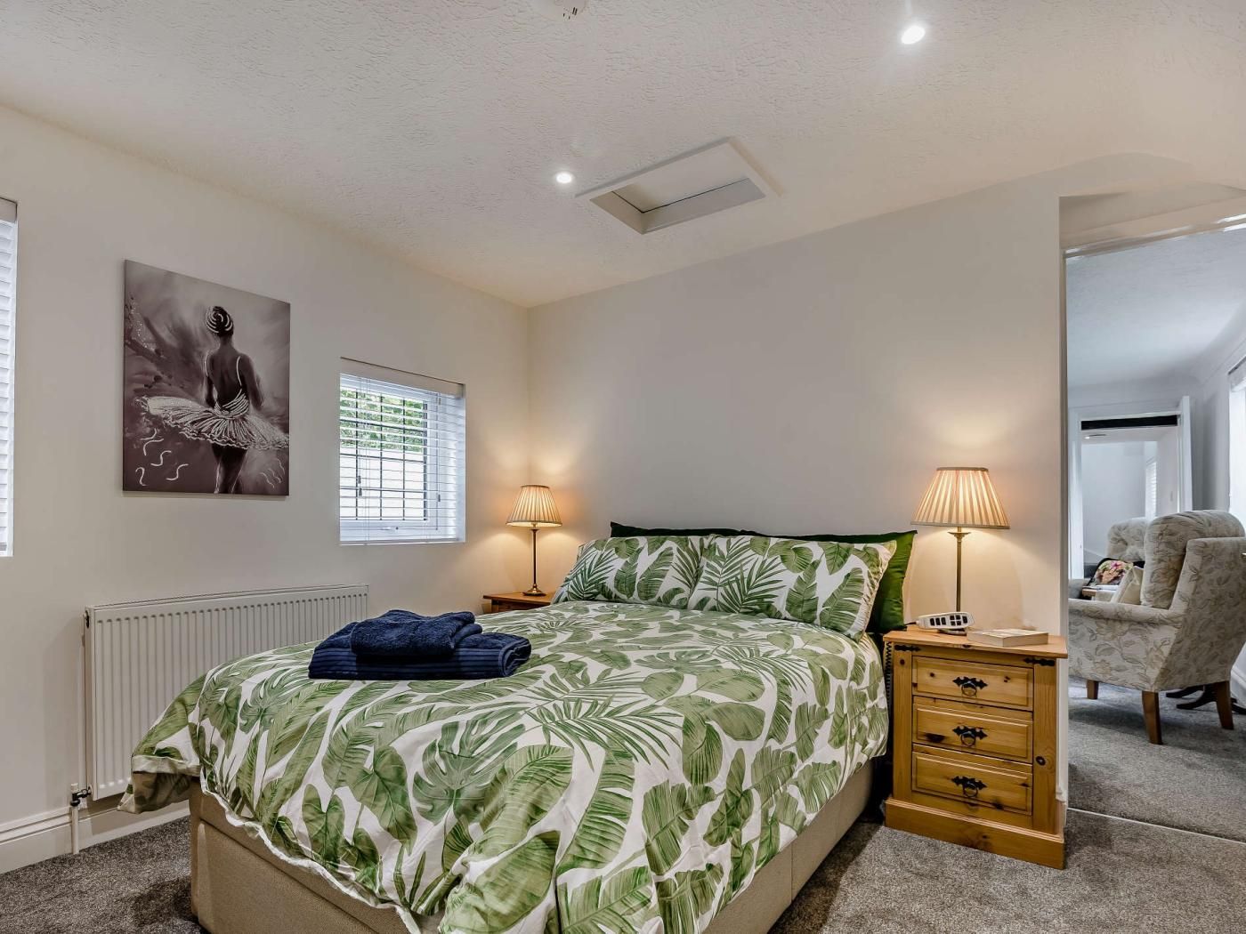 Ein Schlafzimmer mit einem Bett, zwei Nachttischlampen, einem Heizkörper, einem Fenster mit Jalousien, einem Bild an der Wand und einem Sessel in der Ecke.