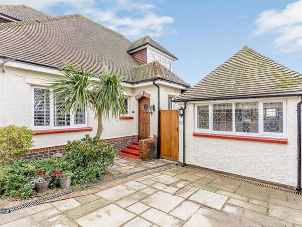 A two-story house with a tiled roof, a front door, and a small garden with a palm tree and potted plants. - 1 Bed in Worthing (oc-80539) (Worthing)