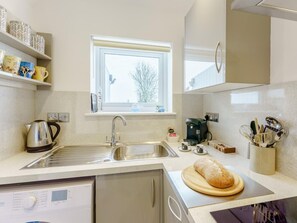 A kitchen with a stainless steel sink, a coffee maker, a bread loaf on a cutting board, and a window with a view of trees. - 1 Bed in Worthing (oc-80539) (Worthing)
