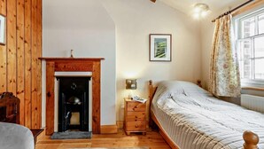A bedroom with a fireplace, a bed, a nightstand, a lamp, a window with curtains, and a wooden ceiling. - 2 Bed in Llanidloes (oc-94088) (Llanidloes)
