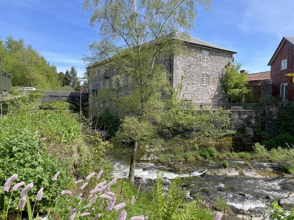 A stone building with a river in front, surrounded by greenery.