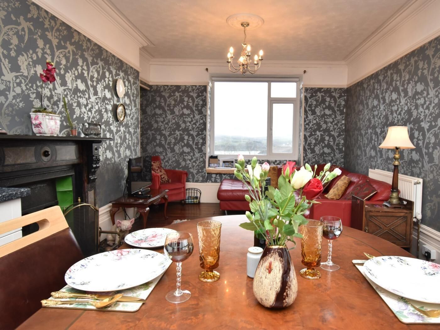 Een eetkamer met een houten tafel voor vier personen, omringd door bloemenbehang, een open haard en een rode bank.