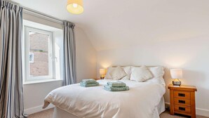 A bedroom with a large bed, two bedside tables with lamps, a window with curtains, and a view of a brick building outside. - 4 Bed in Alnwick (oc-cn155) (Alnmouth)
