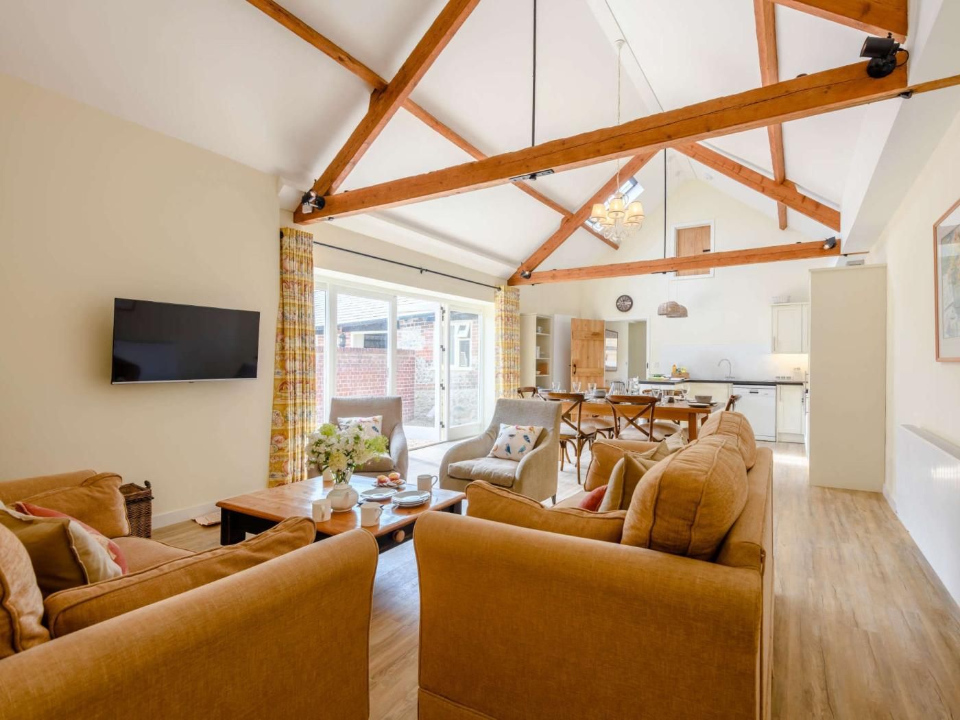 A spacious living room with a high ceiling and exposed wooden beams, a flat-screen TV mounted on the wall, and a dining area with a table and chairs.