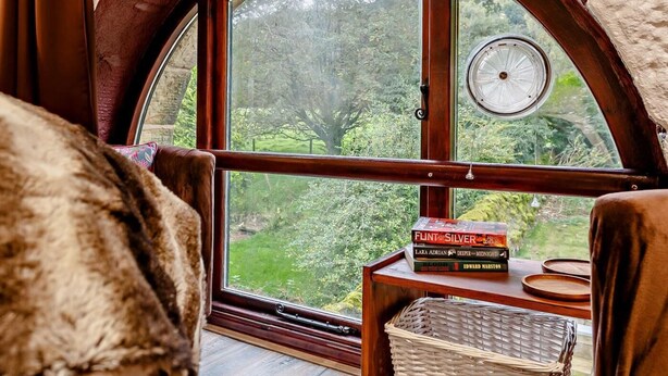 A cozy room with a large arched window, a wooden table with books, and a wicker basket. - 3 Bed in Skipton (oc-93485) (Embsay)