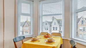 Un coin repas avec une table dressée pour deux personnes, deux chaises et une vue sur les bâtiments par les fenêtres.