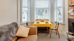 Un coin repas avec une table dressée pour deux personnes, un canapé agrémenté d’un coussin décoratif et un meuble pour la télévision sur lequel sont posés des livres.