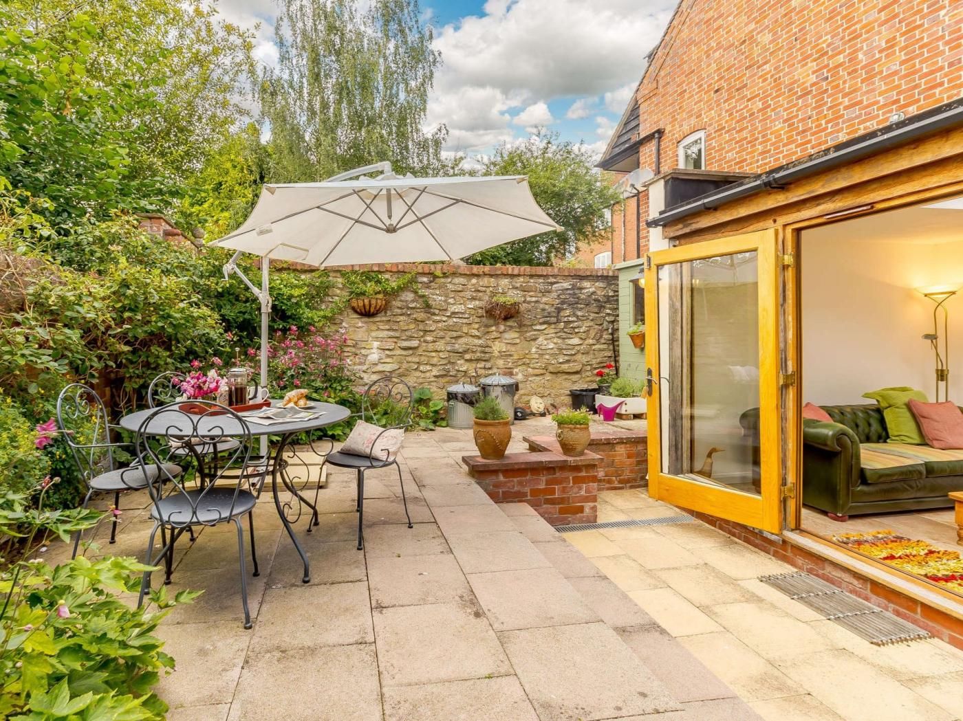 Une terrasse avec une table et des chaises, un mur en pierre et une porte vitrée donnant sur un espace de vie avec un canapé.
