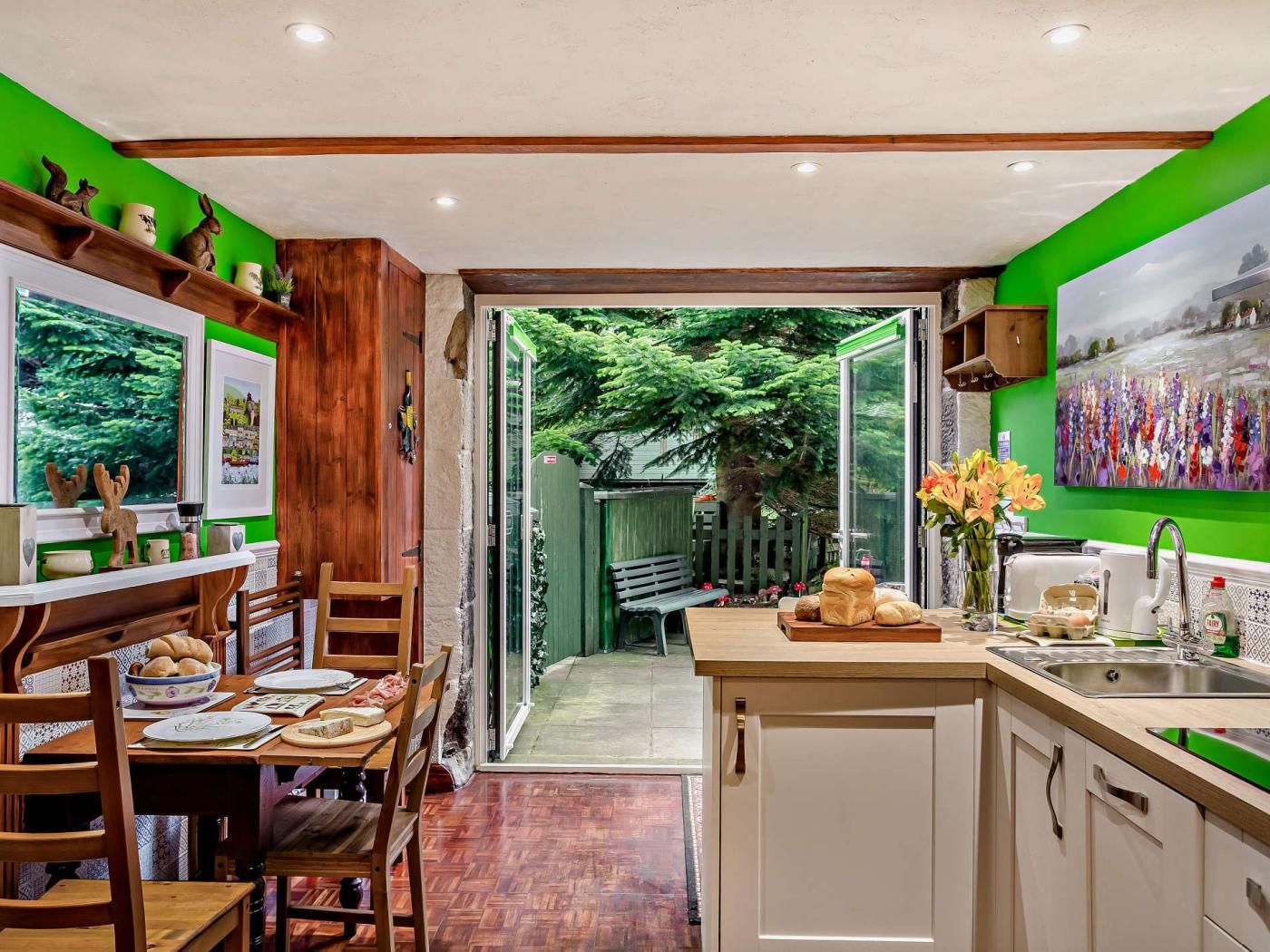 A kitchen with wooden cabinets, a dining area with a table and chairs, and a view of a green outdoor area through an open door.