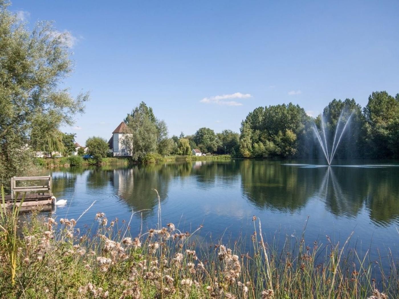A tranquil lake with a fountain, surrounded by trees and a building with a red roof.