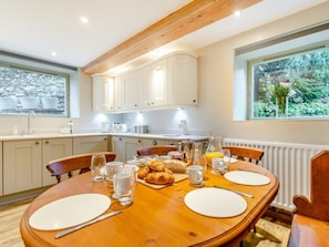 Eine moderne Küche mit weißen Schränken, einem Tisch gedeckt mit Tellern, Gläsern und Croissants, und Blick auf Grün hinter einem großen Fenster.