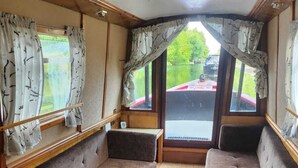 A compact interior with a sofa and bench seating, wooden flooring, and curtains. - Lovely Narrowboat Chiltern Hills and Surrounding. (Hemel Hempstead)