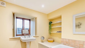 A bathroom with a pedestal sink, a toilet, a bathtub, a window with a mirror, and a shelf with books and a plant. - 3 Bed in Penrith (oc-93379) (Orton)