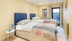 A bedroom with two beds, a small table, a lamp, a window with curtains, and a view of the outside. - 3 Bed in Penrith (oc-93379) (Orton)