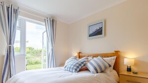 Een slaapkamer met een bed, nachtkastje, lamp, raam met gordijnen en een aan de muur hangend schilderij.