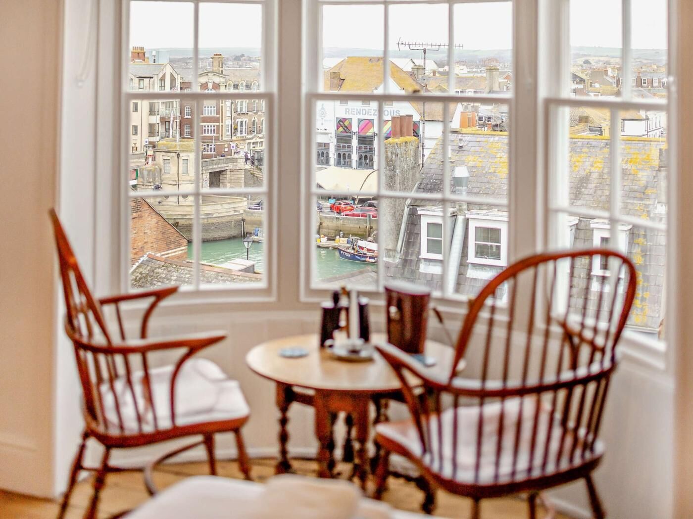 A room with a large window overlooking a harbor, wooden chairs, a small round table, and a view of buildings and boats.