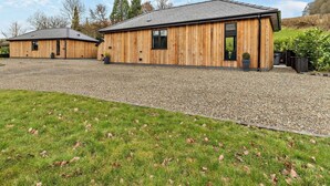 A wooden building with a gravel driveway and a grassy area in front. - 2 Bed in Knighton (oc-94202) (Knighton)