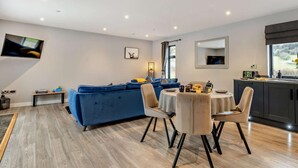 A modern living room with a blue sofa, a dining area with a round table and chairs, a kitchen with dark cabinets, and a large window. - 2 Bed in Knighton (oc-94202) (Knighton)