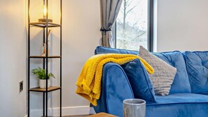 A modern living room with a blue sofa, a yellow throw blanket, a tall floor lamp, a coffee table with a magazine, and a window with curtains. - 2 Bed in Knighton (oc-94202) (Knighton)