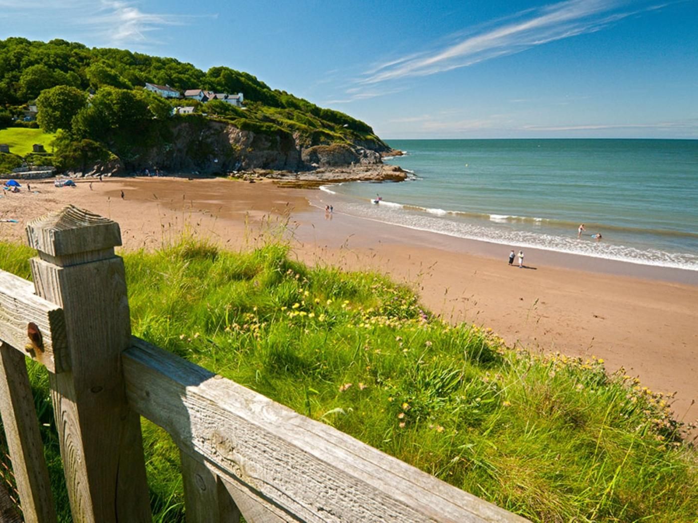 A coastal beach with a wooden fence in the foreground, a grassy area, and a clear sky.