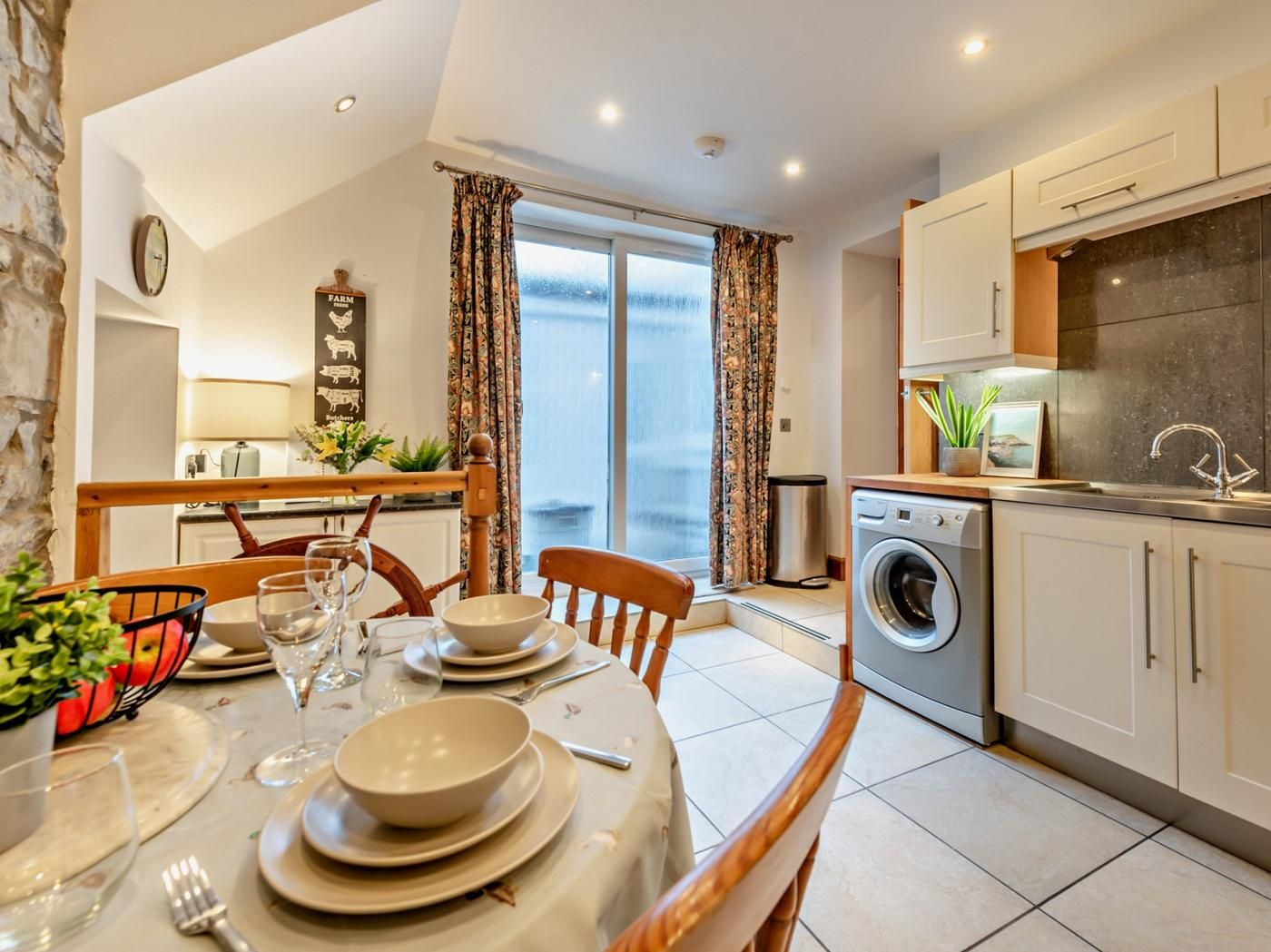 A modern kitchen with a washing machine, a dining area with a table set for four, and a view of the sea through large windows.
