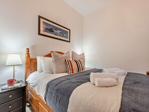 A neatly made bed with a grey and white blanket, a wooden headboard, and a striped pillow. There is a bedside table with a lamp and a red mug. A framed picture hangs on the wall above the bed. - 2 Bed in New Quay (oc-94160) (New Quay)