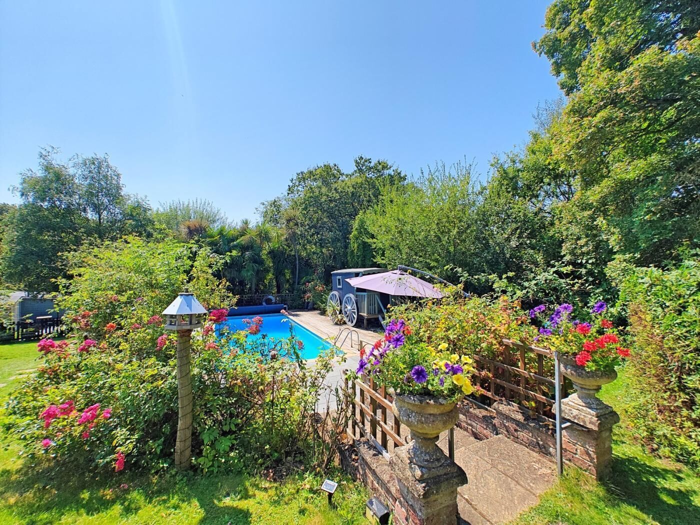 Ein Garten mit Schwimmbad, überdachter Terrasse und üppiger Bepflanzung.