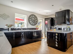 A modern kitchen with black cabinetry, a large wall clock, and a window with a red umbrella view. - 3 Bed in Shanklin (oc-ic180) (Shanklin)
