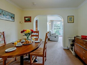 A dining area with a wooden table, chairs, and a vase of flowers. There is a view of a balcony through an archway. - 2 Bed in Ilfracombe (oc-54106) (West Down)
