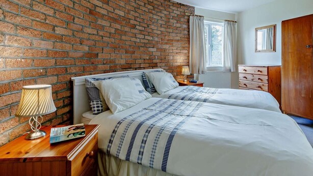Un dormitorio con pared de ladrillo, dos camas, una mesita de noche, una lámpara, un aparador y una ventana con cortinas.