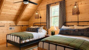 A cozy log cabin bedroom with two beds, a ceiling fan, and a window with checkered curtains.