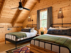 A cozy log cabin bedroom with two beds, a ceiling fan, and a window with checkered curtains.