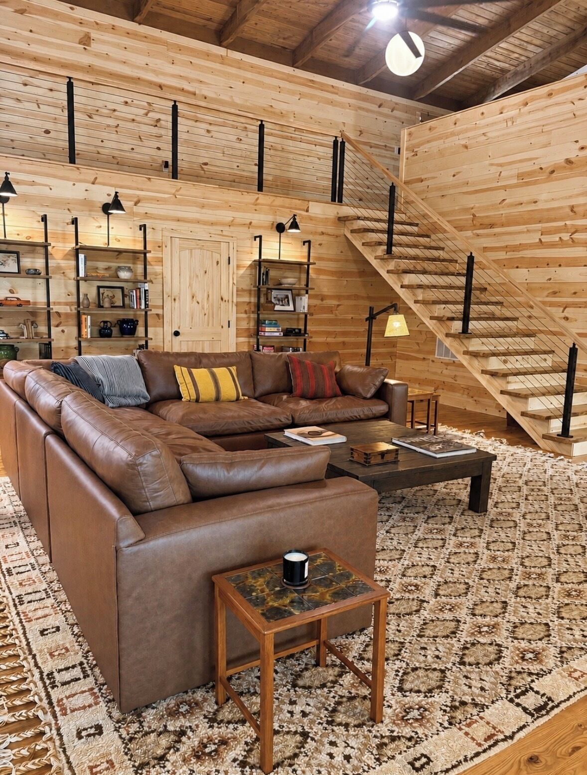 A cozy living room with a brown leather sofa, a coffee table, and a patterned rug. There is a staircase with wooden railings and a wooden ceiling.
