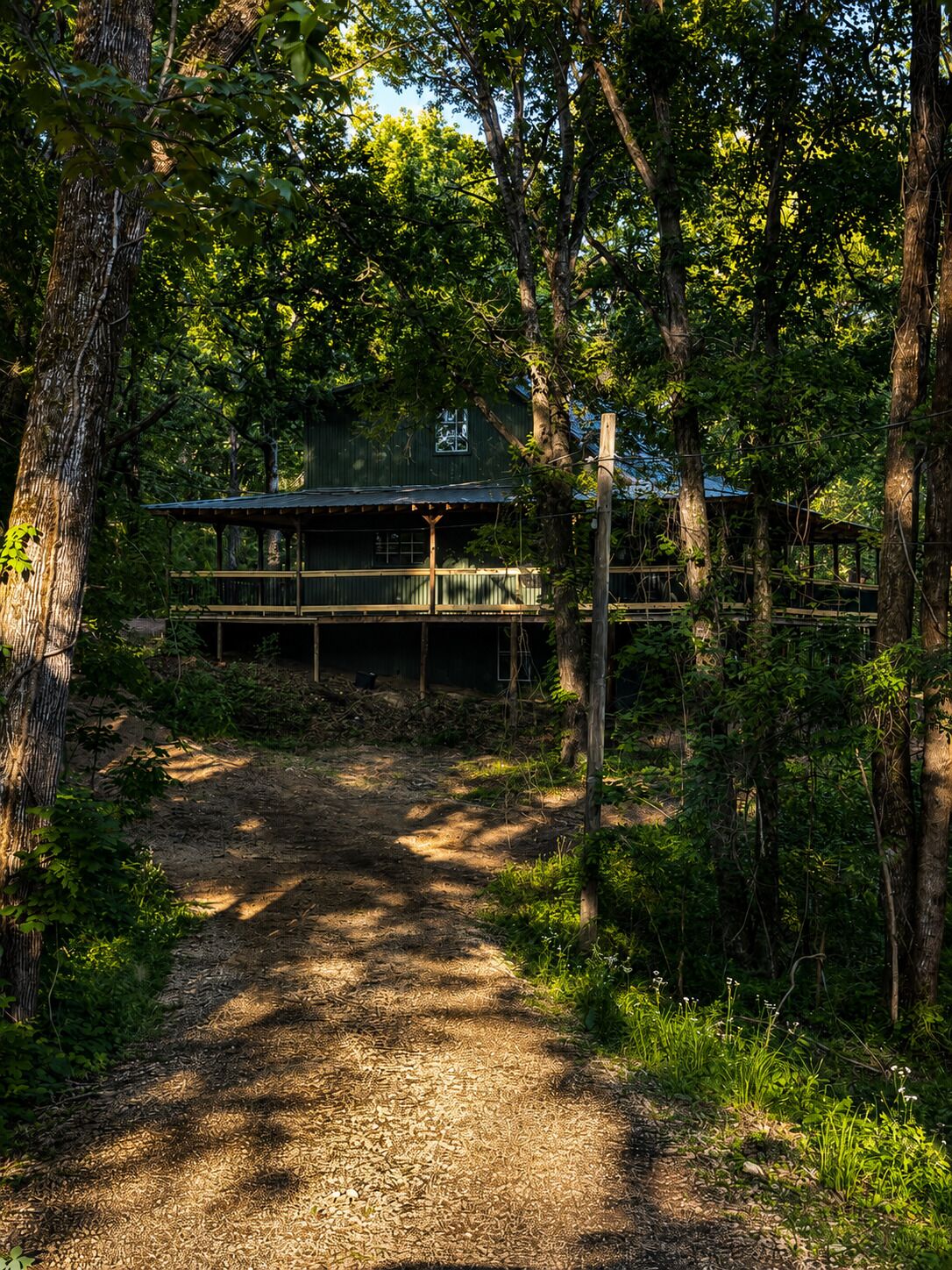 A rustic cabin with a wooden deck and a dirt path leading to it, surrounded by dense forest.
