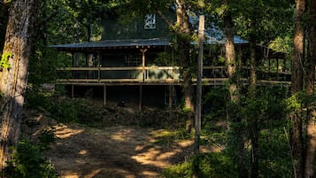 A rustic cabin with a wooden deck and a dirt path leading to it, surrounded by dense forest.