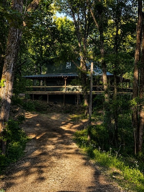 A rustic cabin with a wooden deck and a dirt path leading to it, surrounded by dense forest.